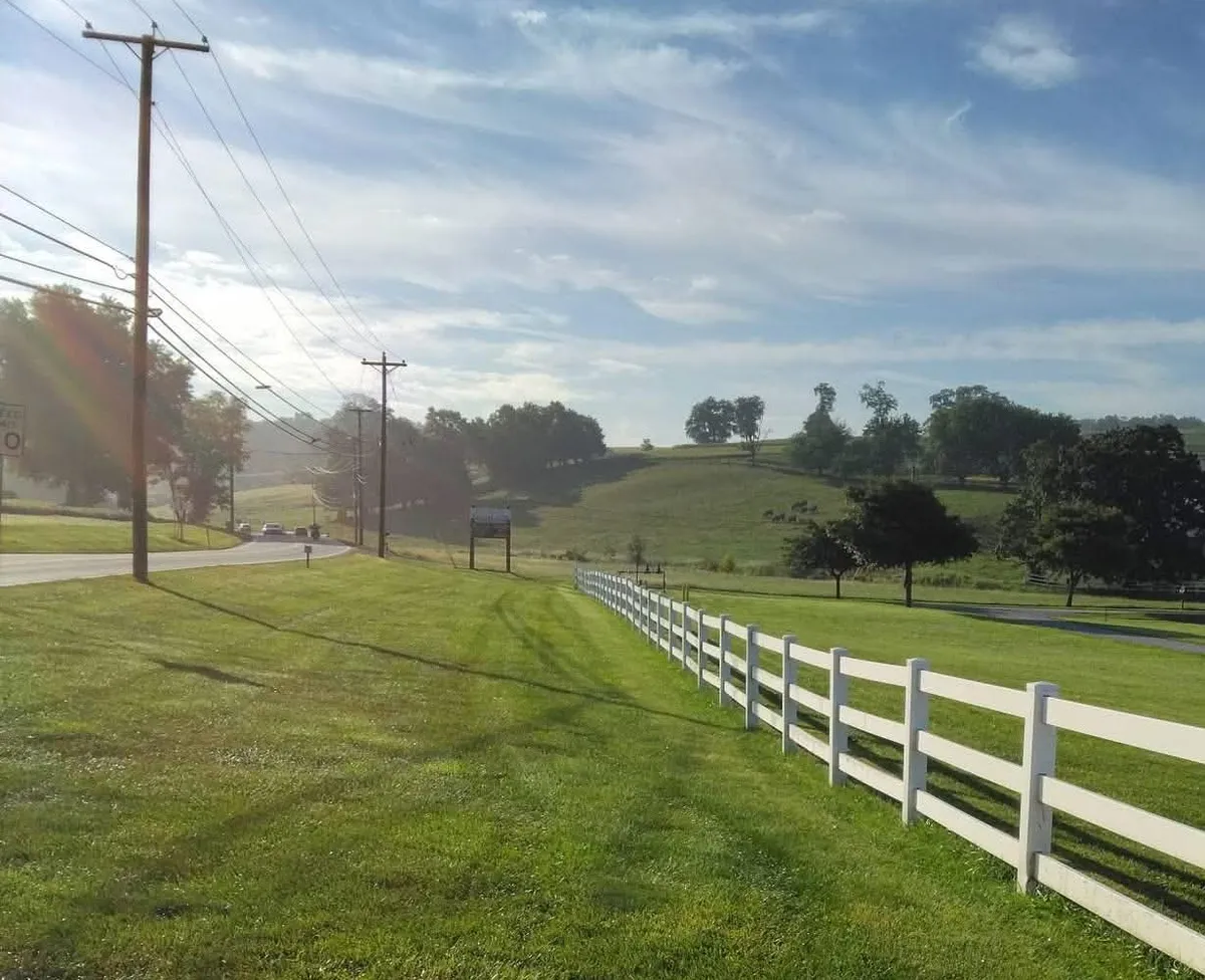 White three-rail fence running along a roadside lawn toward a farm and rolling hills